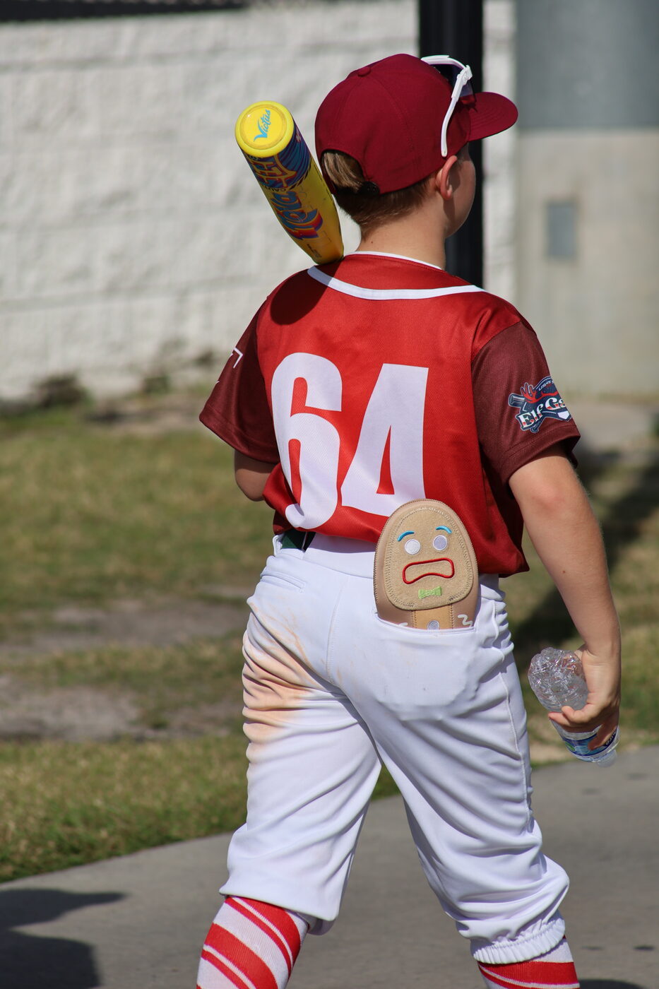 Young player in red #64 jersey carrying a yellow bat, candy-striped socks and a gingerbread-man patch on the back pocket.