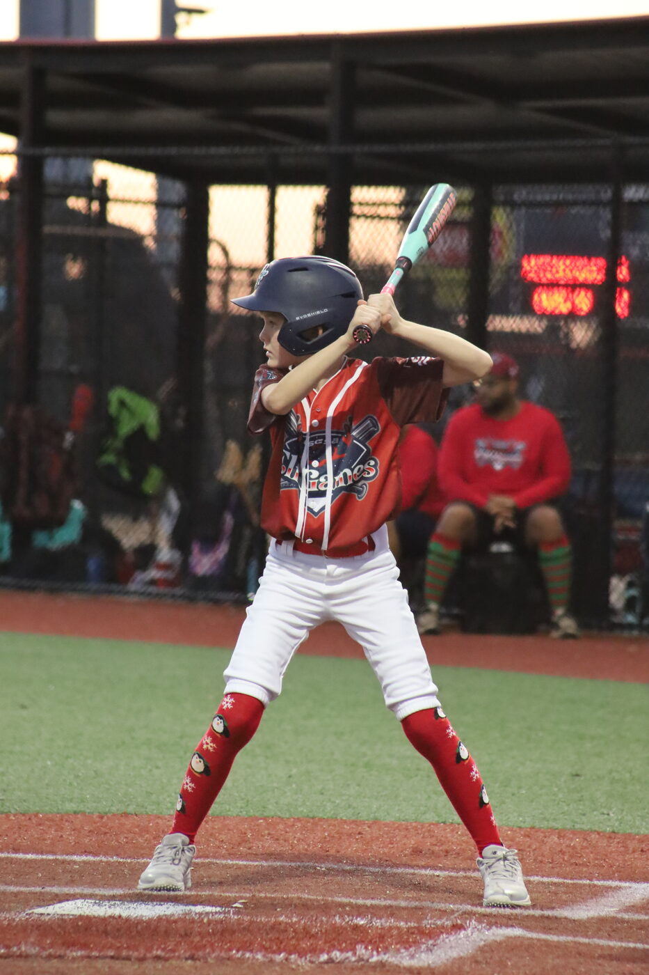 Young batter at the plate in an Elf Games jersey wearing penguin-print Christmas socks.